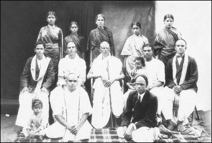 Mahalingam Iyer is sitting dressed in a bow tie, black coat and wearing slippers at extreme right in the middle row. At his back is standing Saradambal, his wife and Meenakshi's mother. Place and date of photo unknown.